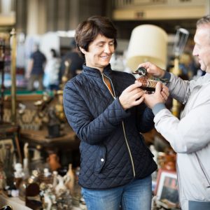 Woman in a quilted navy jacket admires a decorative duck figurine handed by a man in a light jacket inside a cluttered antique shop.