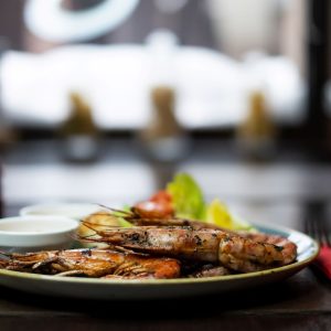 Plate of grilled shrimp with herbs, lemon wedges, and a small bowl of sauce on a dark table; a red napkin sits to the right, with a blurred background.