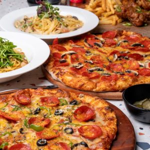 A table of food with two pizzas, pasta, greens, fries, chicken wings, and a bowl of pickles nearby.